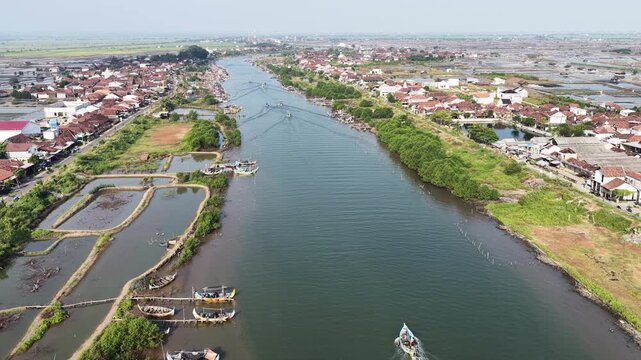 Aerial view of a river with traditional fishing boats moored along the banks and a small boat navigating the water in Jepara, Central Java, Indonesia.