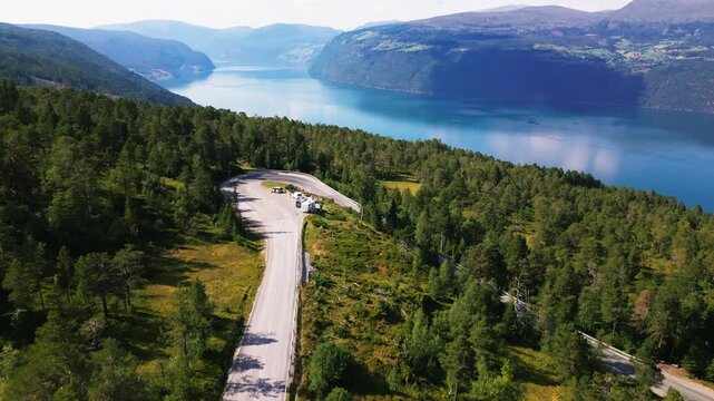 Aerial view of a winding road leading to a scenic parking area overlooking the deep blue waters of the fjord surrounded by lush pine forests in Utvik, Vestland, Norway.