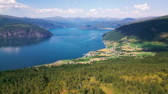 Aerial view of the village of Utvik nestled along the shores of a deep blue fjord, surrounded by lush green fields and towering mountains in Utvik, Vestland, Norway.