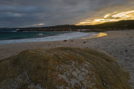 View of Lights Beach with a large lichen-covered rock in the foreground, sandy shore, crashing waves, and golden sunset light in Ocean Beach, Western Australia, Australia.
