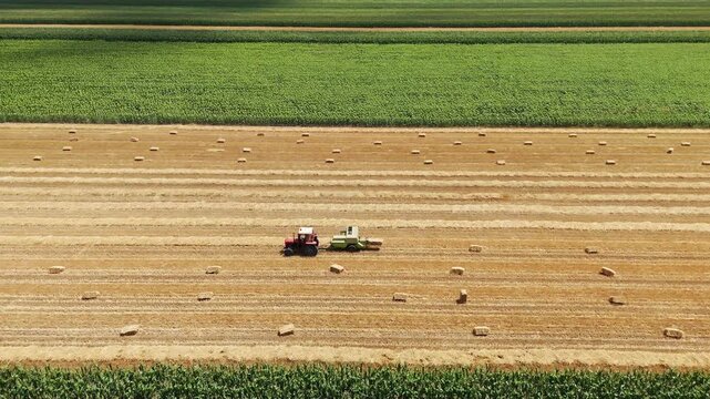 Aerial view of a tractor baling hay in a golden field with rows of bales and green crops in the background in Jazak, Vojvodina, Serbia.