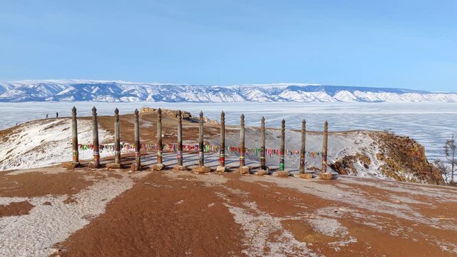 Aerial view of Lake Baikal and wooden ritual poles on the rocky shore of Burkhan Cape with snow-covered landscape under a clear blue sky in Khuzhir, Irkutsk Oblast, Russia.