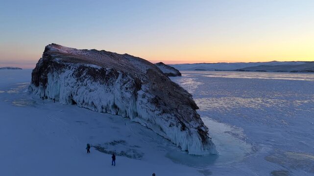 Aerial view of Lake Baikal with people walking on the frozen surface near a rocky island during golden hour in Khuzhir, Irkutsk Oblast, Russia.