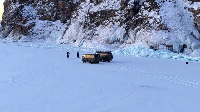 Aerial view of two off-road vehicles parked on the frozen surface of Lake Baikal near snow-covered cliffs with people standing nearby in Khuzhir, Irkutsk Oblast, Russia.