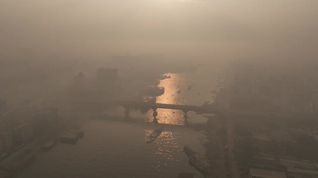Aerial view of the Buriganga River at Sadarghat with boats navigating the hazy water under the golden sunset light in Dhaka, Dhaka Division, Bangladesh.