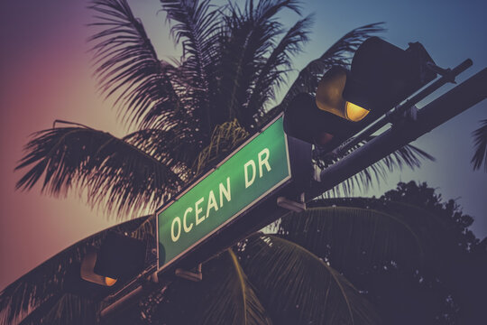 View of the Ocean Drive street sign with traffic lights and palm trees against a colorful sunset sky in Miami Beach, Florida, United States.