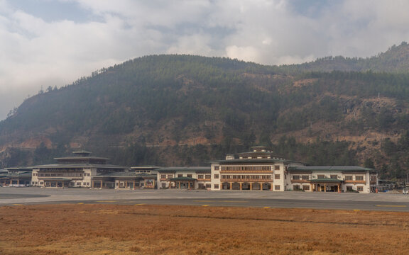 View of Paro International Airport terminal building with its traditional Bhutanese architecture set against a backdrop of forested mountains under a cloudy sky in Paro, Paro, Bhutan.