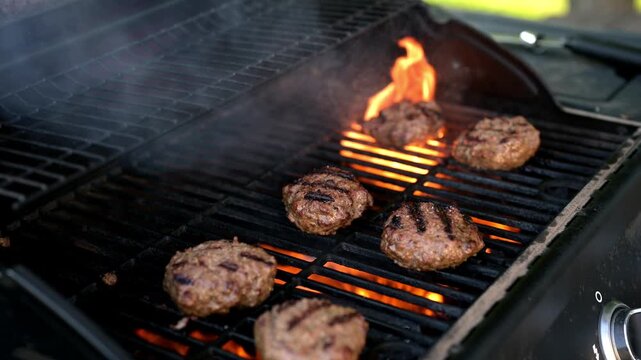4K footage of homemade beef burger patties being flipped as bright flames flare up from a hot outdoor gas grill. This video highlights dramatic flame grilling, backyard barbecues, and summer cookouts