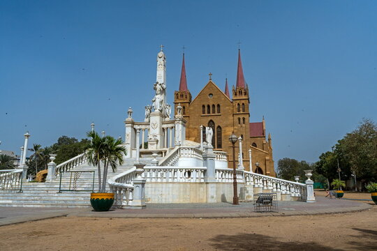 View of St. Patrick's Cathedral with red spires and stone facade, featuring a white marble monument in the foreground under a clear blue sky in Karachi, Sindh, Pakistan.