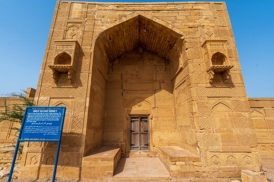 View of the historic sandstone tomb of Isa Khan Tarkhan II at the Makli Necropolis with intricate stone carvings and a blue information sign in Thatta, Sindh, Pakistan.