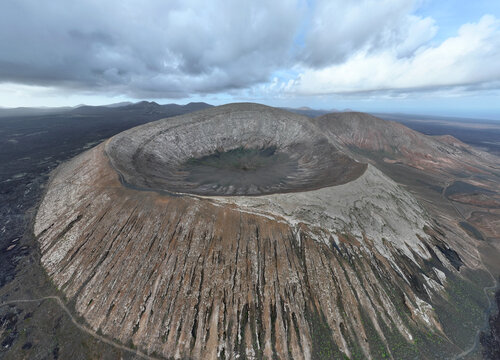 Aerial view of the rugged volcanic crater of Caldera Blanca with dark rocky slopes and deep interior basin under a cloudy sky in Lanzarote.