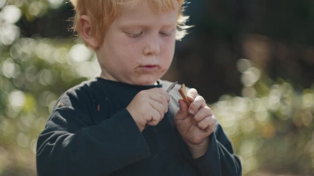 Toddler boy with dirty face cuts wood with little knife and enjoys making something by himself