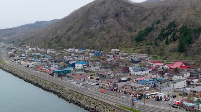 Aerial view of the coastal town of Rausu featuring colorful houses, a road along the shoreline, and mountains in the background in Rausu, Hokkaido, Japan.