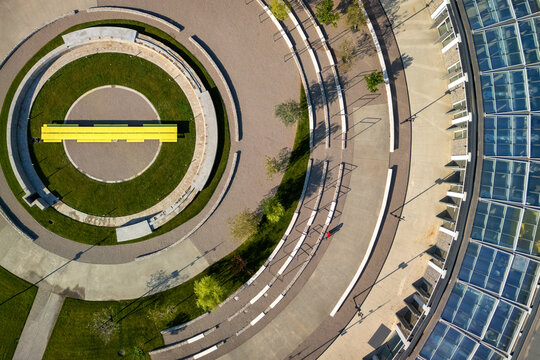 Aerial view of a modern circular plaza with a bright yellow bench, concentric stone pathways, green lawns, and a large glass-roofed building in Belgrade, City of Belgrade, Serbia.