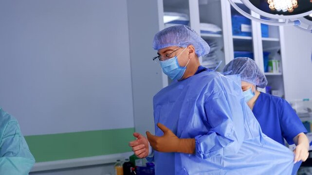 Male doctor is being helped to get dressed for the surgery. Two nurses help the surgeon to put on robe and gloves for the work at operation.