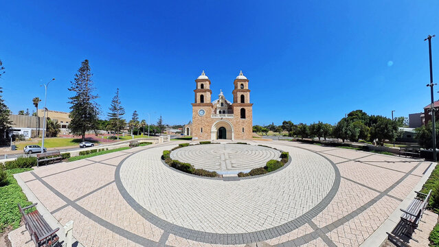 View of St Francis Xavier's Cathedral with its stone facade and twin towers, featuring a circular labyrinth in the plaza under a clear blue sky in Geraldton, Western Australia, Australia.
