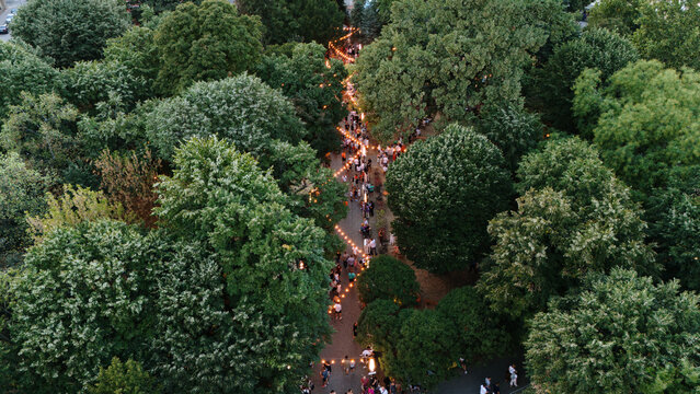 Aerial view of a park pathway illuminated by string lights with people walking among lush green trees in Sremska Mitrovica, Serbia.
