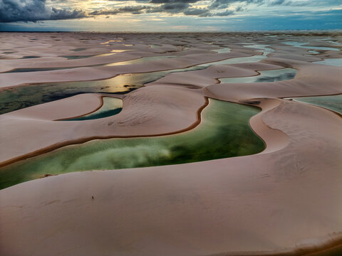 Aerial view of Lagoa Bonita with its emerald green lagoons nestled among vast white sand dunes under a dramatic sunset sky in Barreirinhas, Maranhao, Brazil.