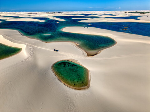Aerial view of Parque Nacional dos Lencois Maranhenses featuring white sand dunes and turquoise lagoons with a vehicle driving on the sand in MA, Brazil.