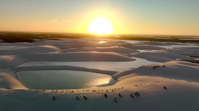 Aerial view of Parque Nacional dos Lencois Maranhenses featuring vast white sand dunes, crystal clear lagoons, and off-road vehicles at sunset in MA, Brazil.