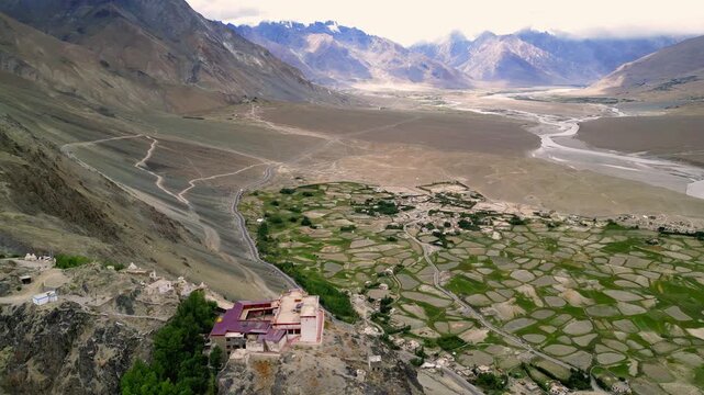 Aerial view of Zanskar Valley monastery perched on a rocky cliff with green terraced fields and a winding river in Zanskar, Ladakh, India.