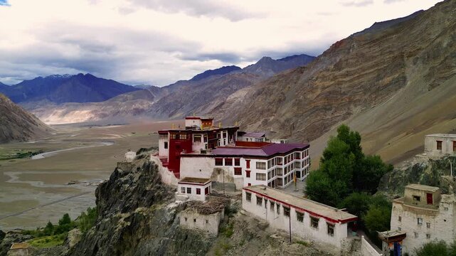 Aerial view of Karsha Monastery perched on a rocky cliff overlooking the Zanskar valley with rugged mountains and a winding river under a cloudy sky in Zanskar, Ladakh, India.