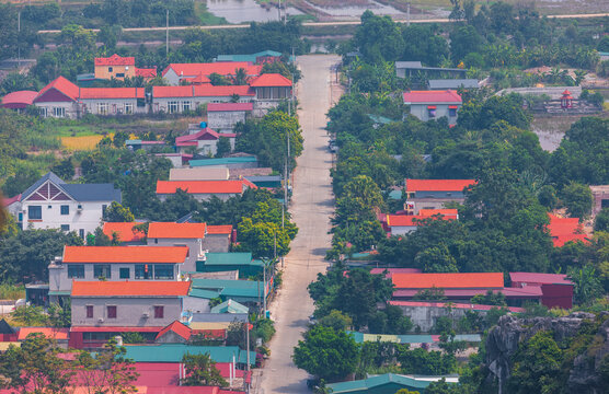 Aerial view of Tam Coc - Bich Dong area featuring a straight road flanked by houses with red roofs, lush green trees, and limestone karst landscape in Nam Hoa Lu, Ninh Binh, Vietnam.