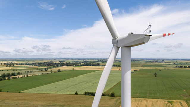 Aerial view of a wind turbine standing in the middle of lush green and golden agricultural fields under a bright blue sky in Nowy Dwor Gdanski, Pomeranian Voivodeship, Poland.