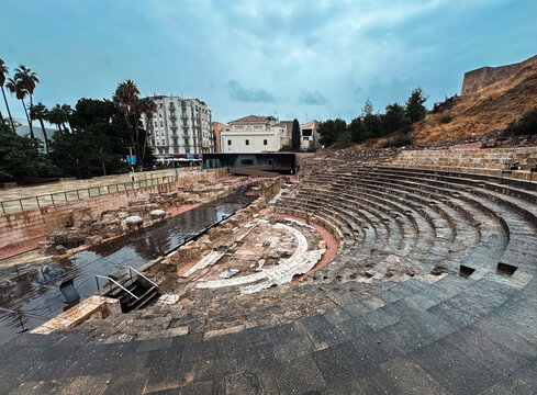 Malaga, Spain - 28 October 2025: View of the Roman Theatre of Malaga with its ancient stone seating, stage area, and surrounding historic urban architecture under a cloudy sky.