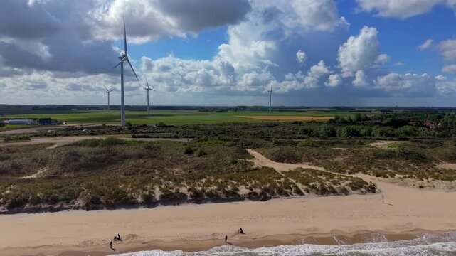 Aerial view video clip of sandy beach, marram grass dunes, wind turbines, agricultural fields, and dynamic sky with cumulus clouds in Domburg, Zeeland, Netherlands on a sunny day