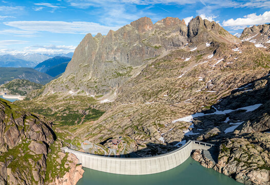 Aerial view of Barrage d'Emosson, a concrete dam in the rugged alpine landscape with rocky slopes, snow patches, and a turquoise reservoir in Finhaut, Valais, Switzerland.