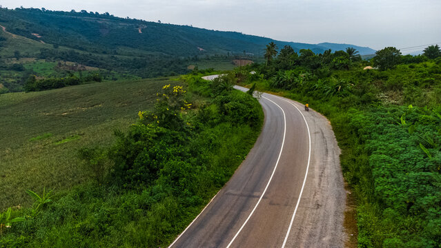 Aerial view of a winding road cutting through lush green hills in the Pokrom area, with dense vegetation and a lone motorcycle on the asphalt in Aburi, Eastern Region, Ghana.