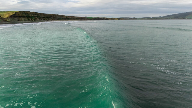 Aerial view of Gemstone Beach with turquoise waves rolling onto the shore, dark coastal cliffs, and a cloudy sky in Orepuki, Southland Region, New Zealand.