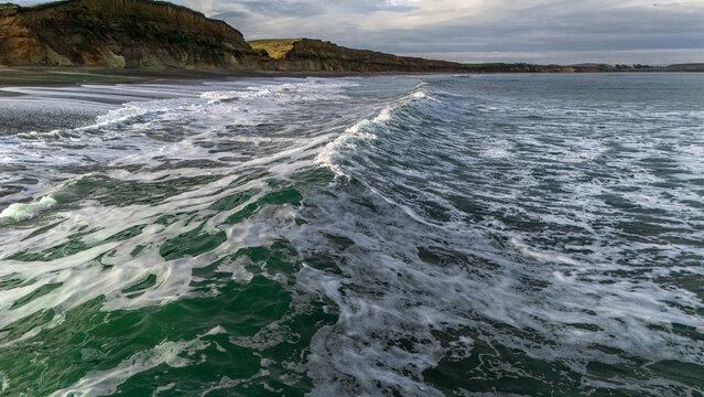 Aerial view of Gemstone Beach with crashing waves, dark sand, and coastal cliffs under a cloudy sky in Orepuki, Southland Region, New Zealand.