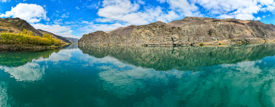 Aerial view of the turquoise waters of Lake Dunstan reflecting the rugged rocky hills and golden willow trees under a bright blue sky in Cromwell, Otago Region, New Zealand.