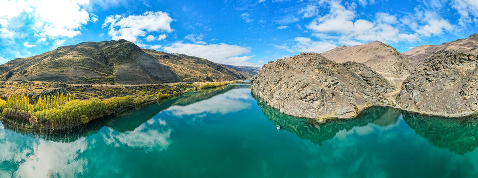 Aerial view of the turquoise waters of Lake Dunstan winding through arid, rocky hills with yellow autumn trees along the banks in Cromwell, Otago Region, New Zealand.