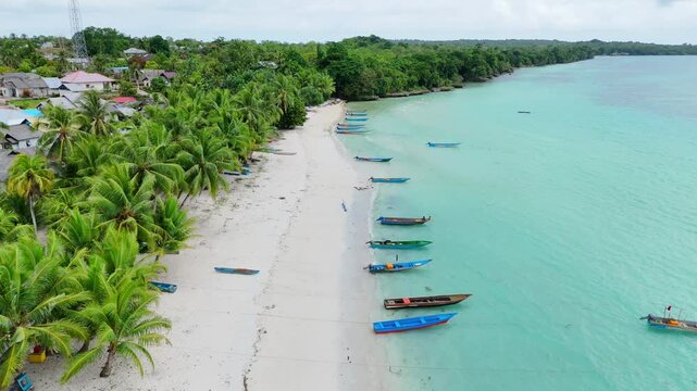 Aerial view of Pantai Ohoidertutu Ngur Mun Vat Wahan with white sand beach, palm trees, and traditional wooden boats in the turquoise water of Kei Kecil Barat, Maluku, Indonesia.