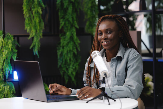 Smiling african female young adult podcaster with laptop and microphone in indoor studio setting, online video streaming broadcast for subscribers videovlog