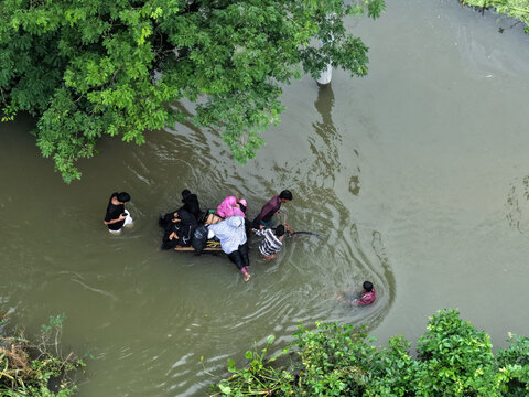 Banshkhali Upazila, Chittagong Division, Bangladesh - 10 July 2026: Aerial view of people wading through floodwaters, pushing a makeshift raft through murky water near lush trees.