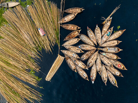 Aerial view of Buriganga River featuring a cluster of wooden boats arranged in a circular pattern alongside stacks of bamboo poles in the water in Dhaka Division, Bangladesh.
