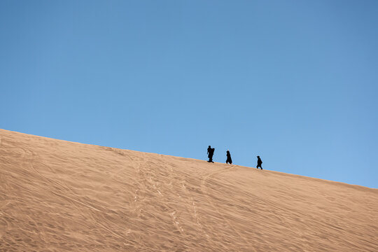 View of people sandboarding in Death Valley, walking up a large sand dune under a clear blue sky in San Pedro de Atacama, Antofagasta, Chile.