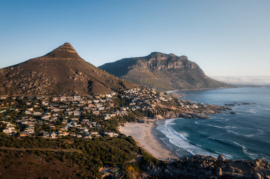 Aerial view of Llandudno Beach with sandy shore, residential houses on the hillside, and dramatic peaks of the Twelve Apostles mountain range in Cape Town, Western Cape, South Africa.