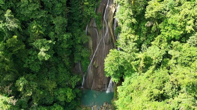 Aerial view of Air Terjun Taeno waterfall cascading into a turquoise pool surrounded by dense tropical rainforest in Teluk Ambon, Maluku, Indonesia.