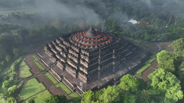 Aerial view of Borobudur Temple surrounded by lush green trees and misty atmosphere in Kecamatan Borobudur, Jawa Tengah, Indonesia.