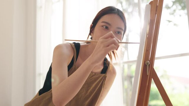 Creative young Asian woman artist painting on canvas in bright home studio. Relaxed female hobbyist enjoying peaceful indoor leisure time, expressing imagination and passion for art.