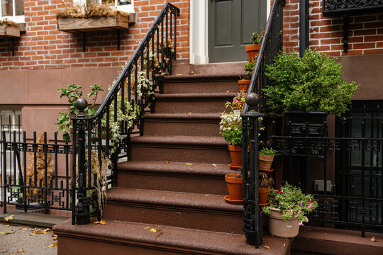 Close up view of brownstone stairs with black iron railings and multiple potted plants in a New York City neighborhood. Brick facade and decorative details highlight classic urban residential