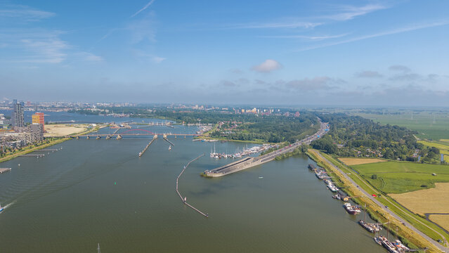 Aerial view of the IJ waterway with the Schellingwouderbrug, modern buildings, boats moored along the bank, and green fields under a blue sky in Amsterdam, North Holland, Netherlands.