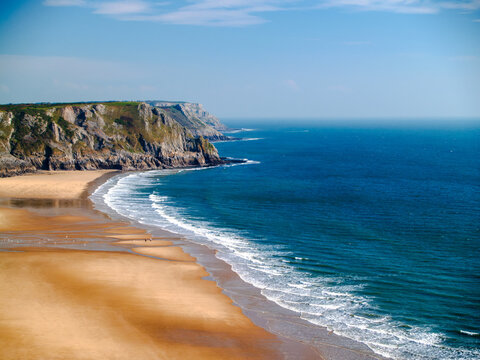 Aerial view over Three Cliffs Bay, golden sand beach curving beneath limestone headland, turquoise sea, Gower Peninsula, Wales.