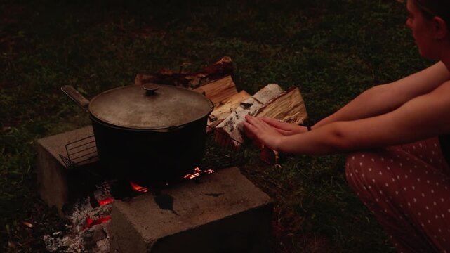 A young woman warming her hands near a large pot cooking over an open campfire outdoors in the evening. pyrotherapy concept. campfire people cooking