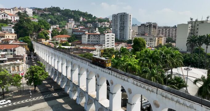 Aerial view of the historic Arcos da Lapa aqueduct with a yellow tram crossing the arches, surrounded by lush palm trees and urban buildings in Rio de Janeiro, State of Rio de Janeiro, Brazil.